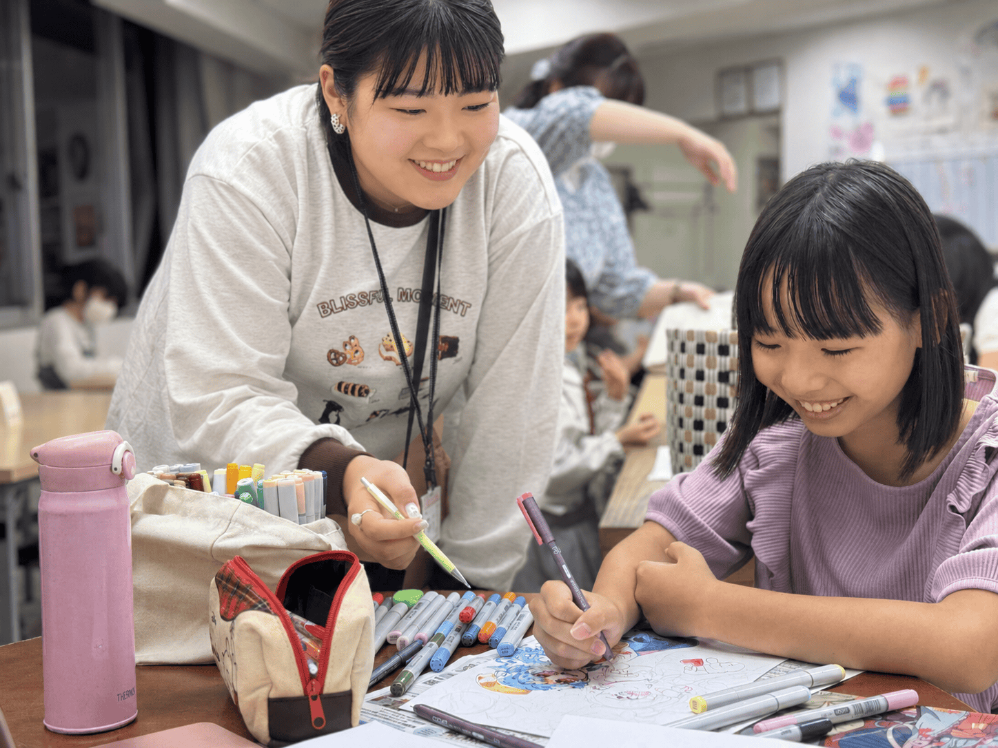 講師と一緒に、机で作品を描き進める笑顔の生徒
